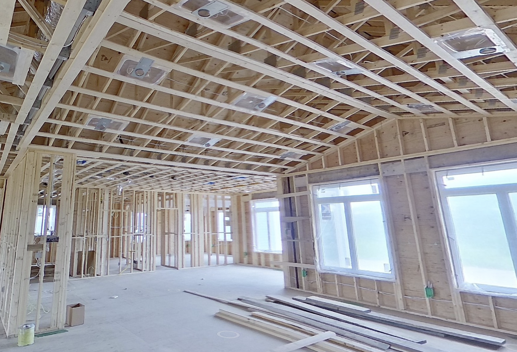 Unfinished home interior with exposed wood framing, ceiling trusses, installed windows, recessed lighting, and lumber on the concrete floor