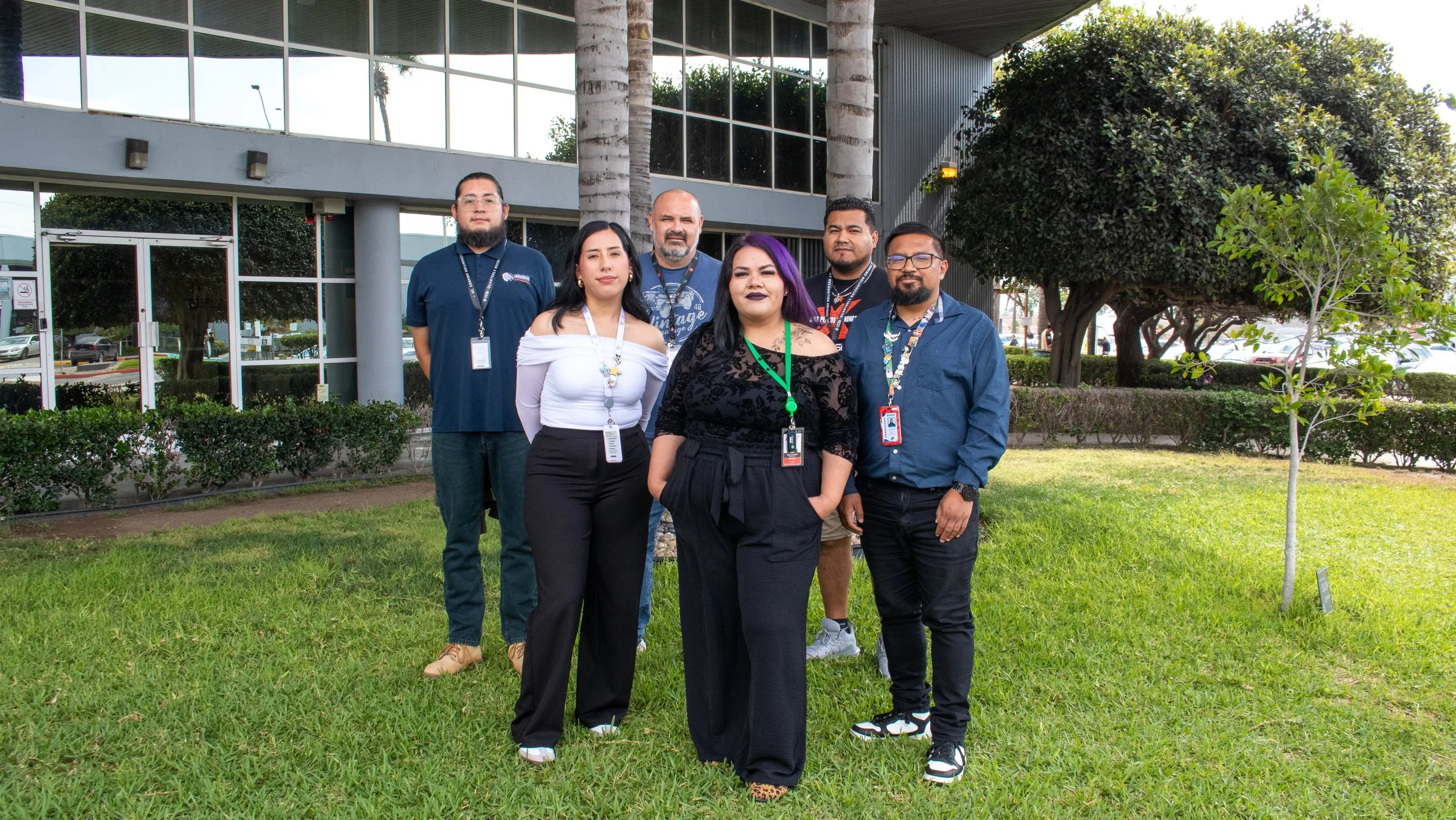 Group of six people standing outside in front of a modern building with glass windows, trees, and bushes, posing for a photo.
