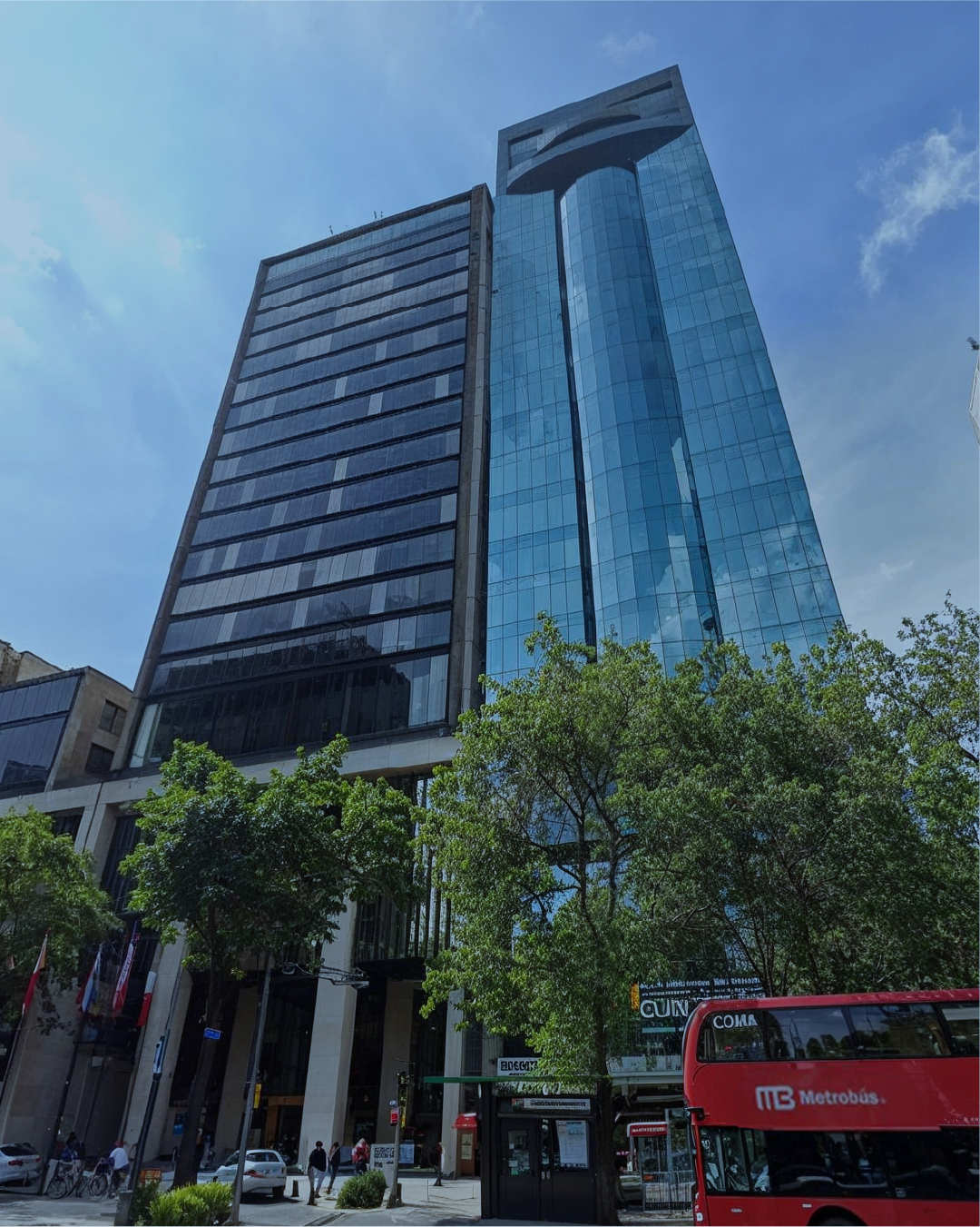Tall modern glass office skyscraper with blue reflective windows, surrounded by trees and a city street with people, cars, and a red double-decker bus.