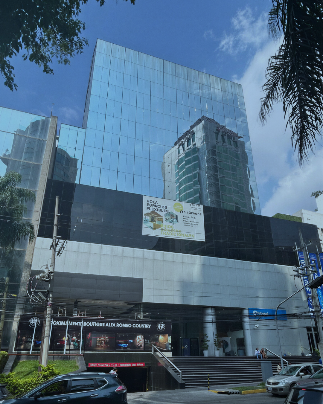 Modern glass office building with reflective windows, palm trees, cars parked in front, and a sign for a boutique Alfa Romeo car dealership.