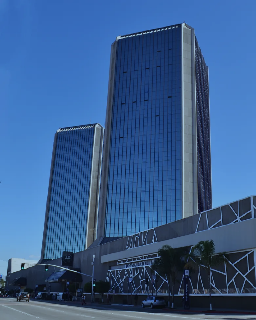 Two tall modern glass skyscrapers with blue reflective windows against a clear blue sky, with the lower floor labeled 'Grand Hotel' in an urban setting.