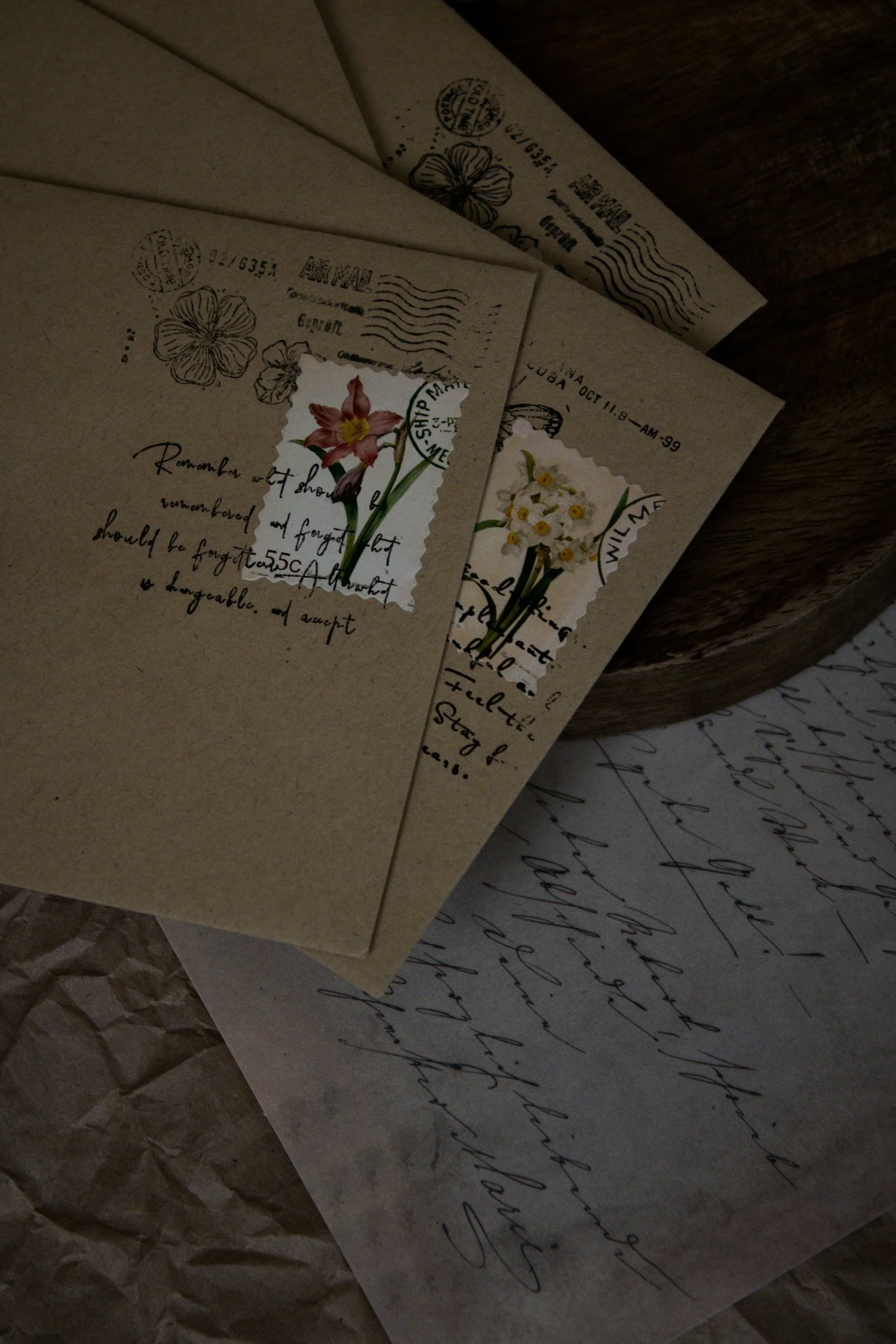 Several pieces of vintage-style paper and a card with handwritten cursive text, floral illustrations, and postage stamp designs, placed on a crumpled brown paper surface.
