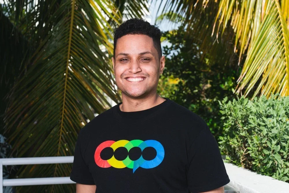 A smiling man with short curly hair wearing a black T-shirt with rainbow-colored circles and speech bubble design, standing outdoors with palm trees and greenery in the background.