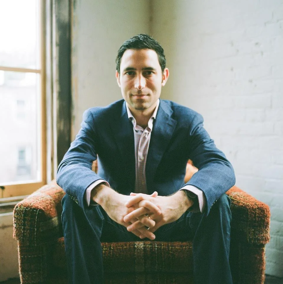A man wearing a dark blazer and a checkered shirt sitting on a red and orange patterned couch, with a window and a white brick wall in the background.
