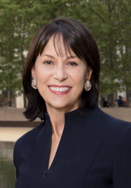 A woman with short dark hair, wearing a navy blue top and large earrings, smiling outdoors with trees and a park in the background.
