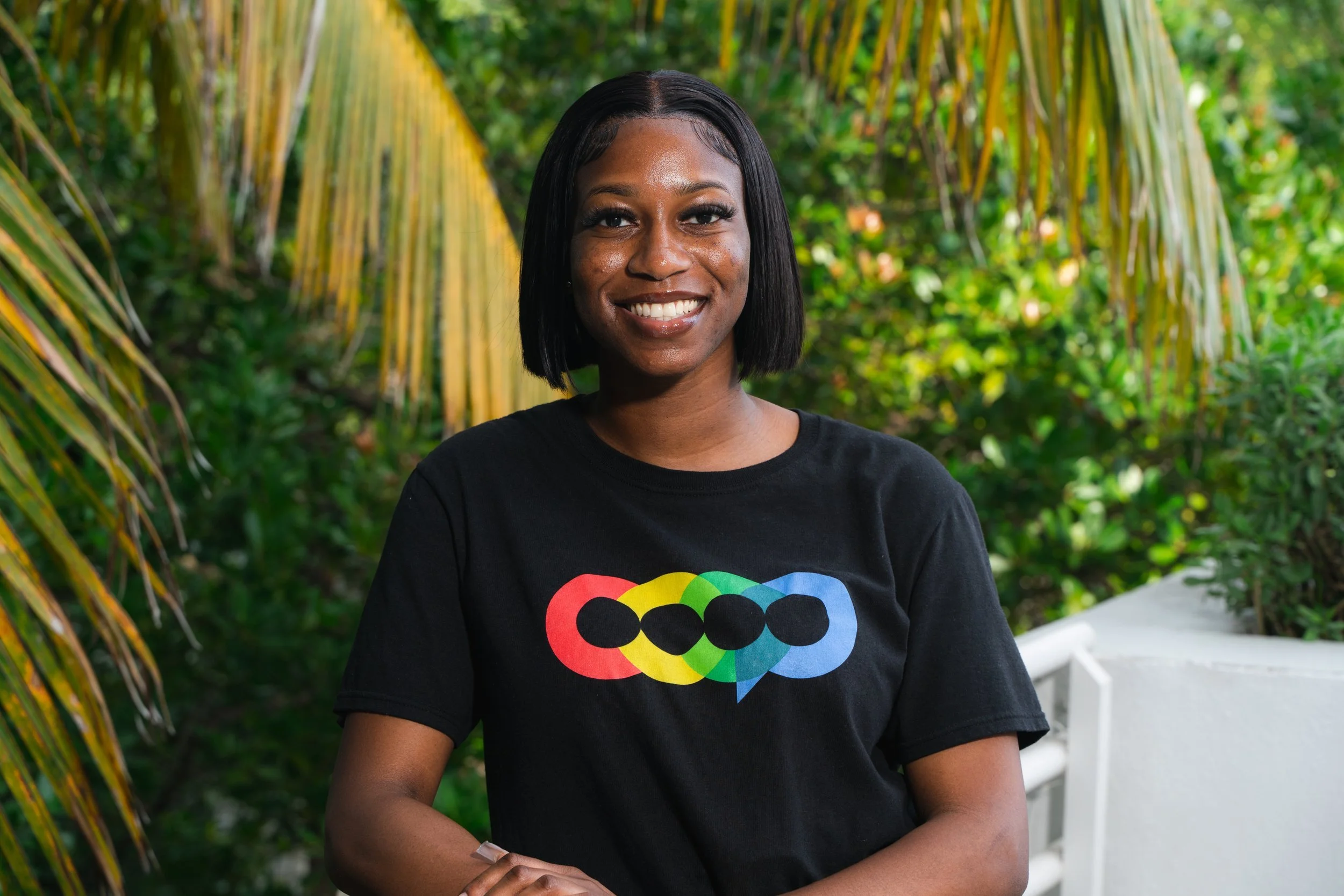 A smiling woman with dark skin and shoulder-length black hair standing outdoors in front of green foliage and yellow palm leaves, wearing a black T-shirt with a colorful graphic of overlapping circles.