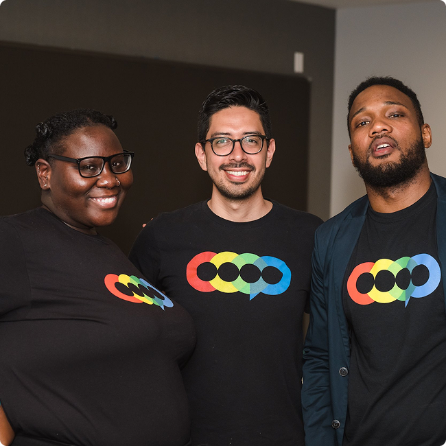 Three smiling people wearing black t-shirts with a colorful rainbow-colored logo, standing close together indoors.