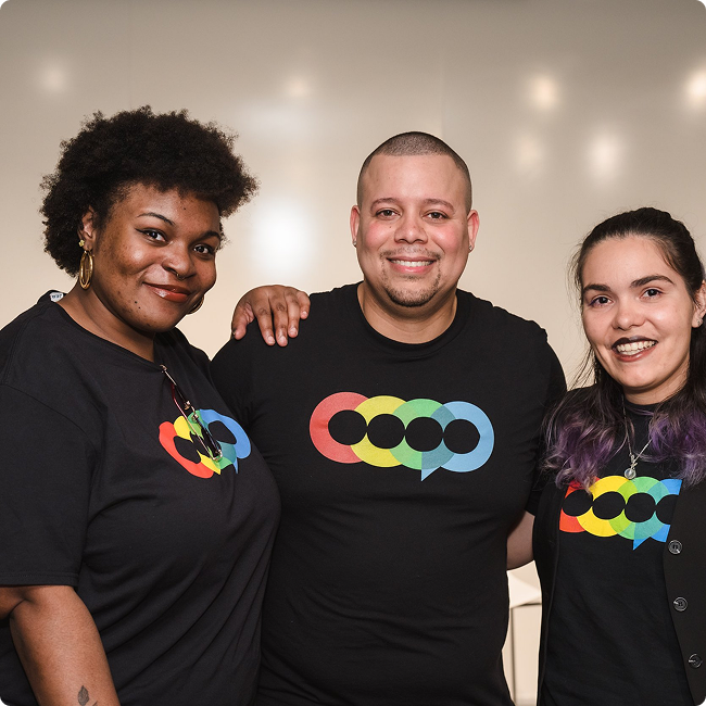 Three diverse people smiling and standing together indoors, wearing matching black T-shirts with colorful speech bubble designs.