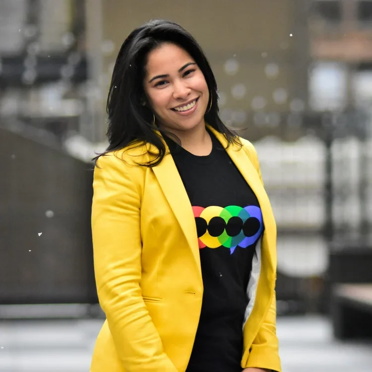 A young woman with black hair, smiling, wearing a yellow blazer and a black t-shirt with a colorful, circular graphic design. She is standing outdoors with a blurred background.
