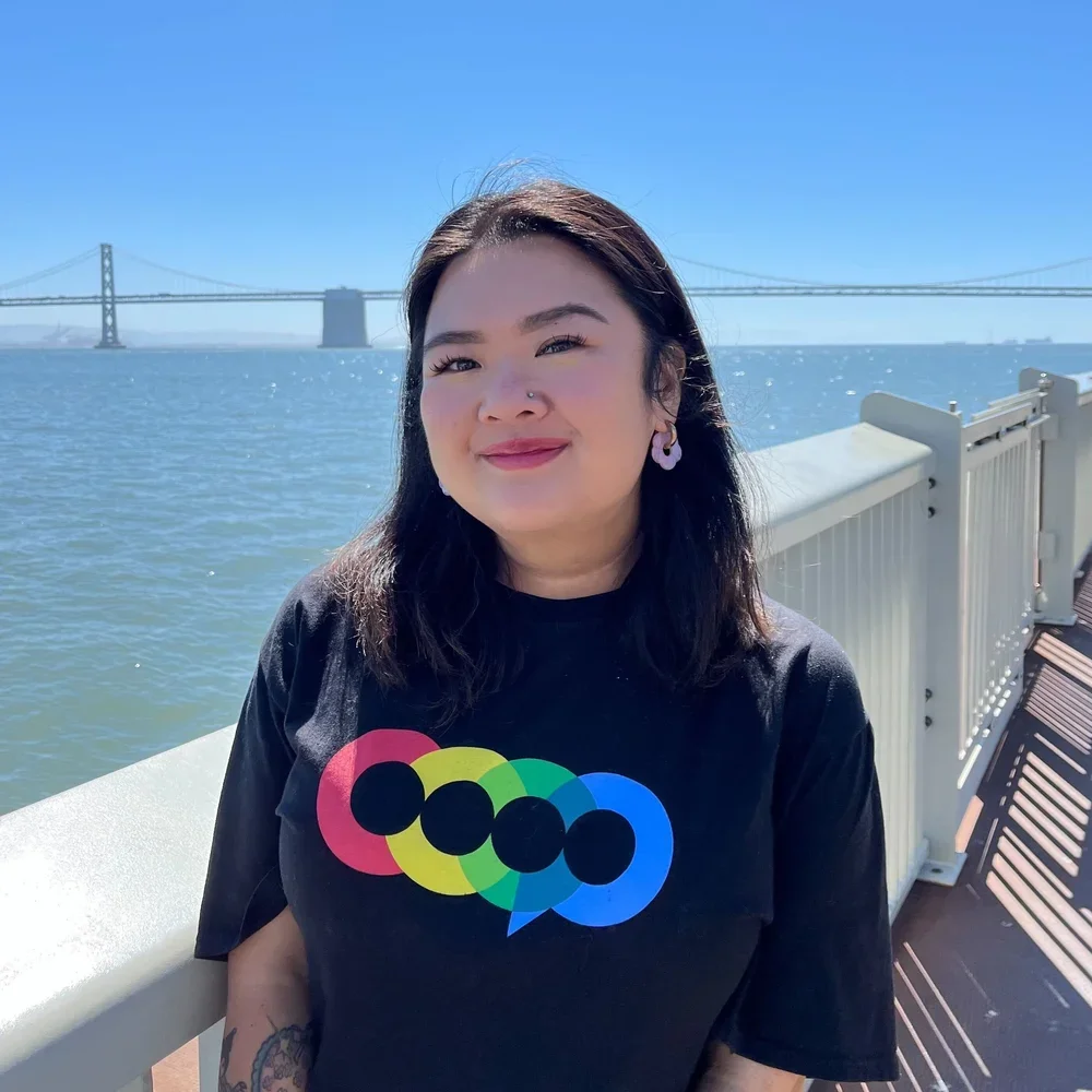 A woman standing outdoors by a railing with a water body and a bridge in the background, smiling and squinting at the camera, wearing a black T-shirt with colorful overlapping circle patterns and hoop earrings.