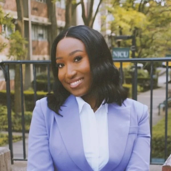Smiling young woman with shoulder-length black hair in a lavender blazer and white blouse, standing outdoors in front of a metal railing and trees.