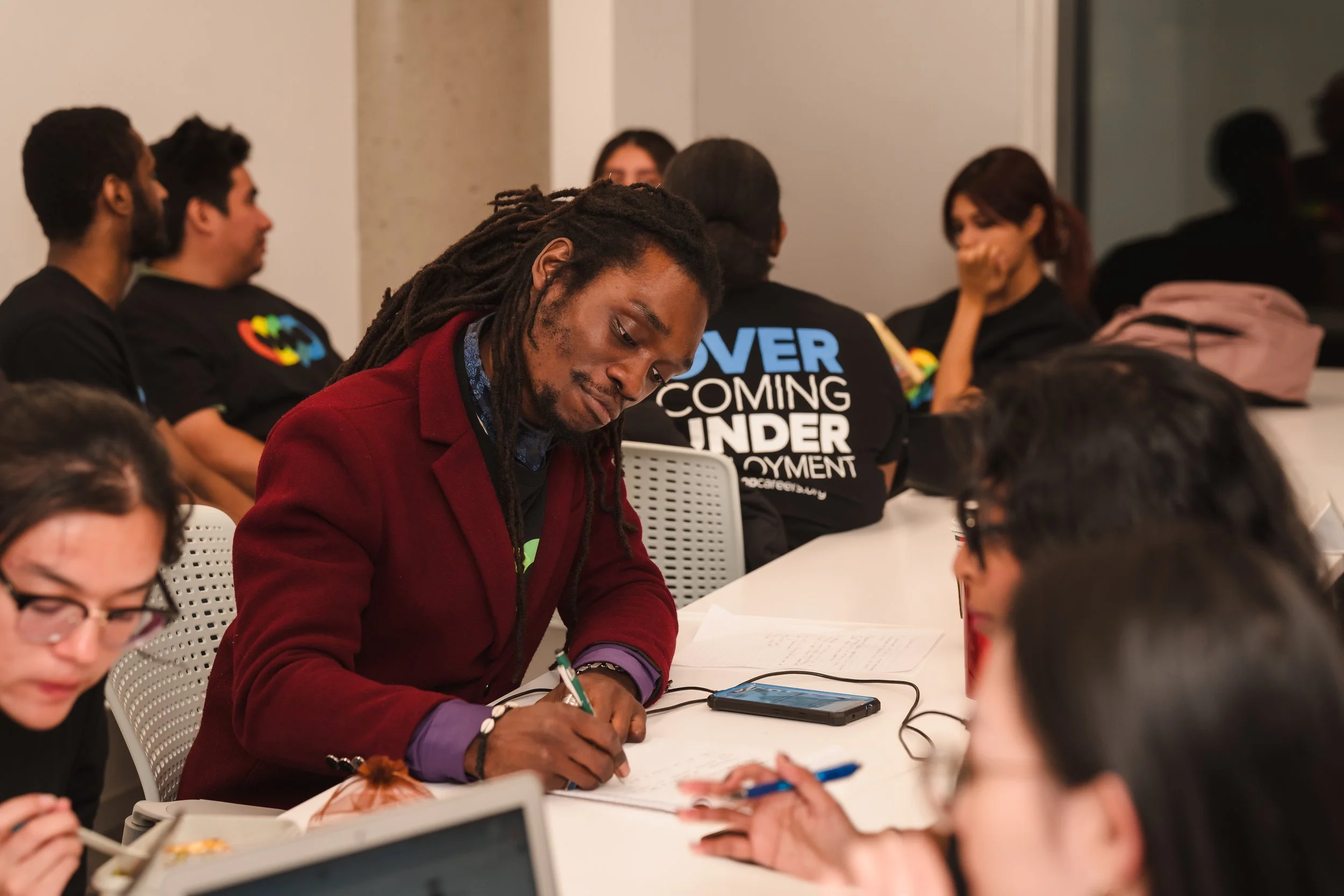 A group of diverse young adults sitting at a long table during a meeting or workshop, with some taking notes and using electronic devices.