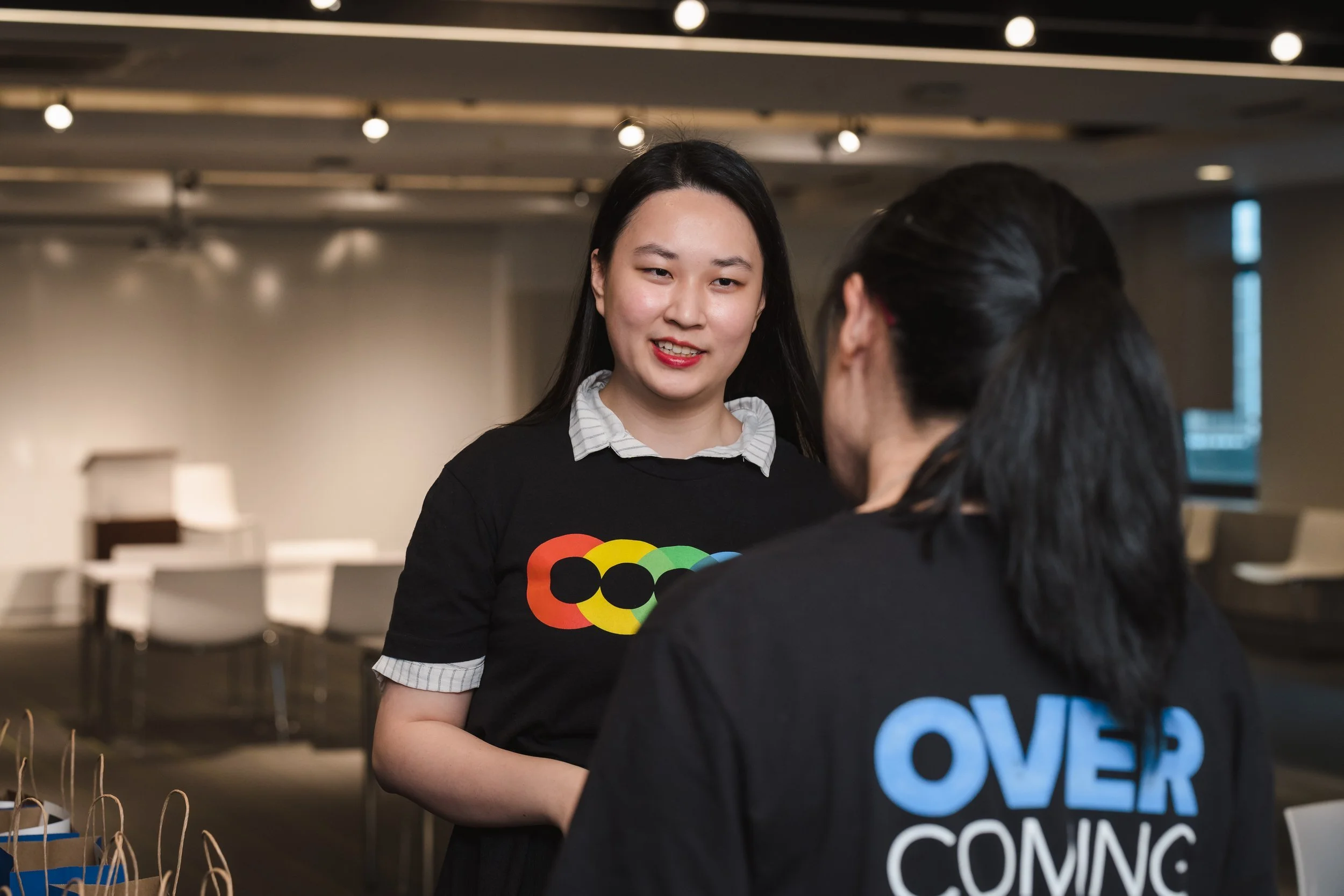 Two women having a conversation in a well-lit room, one woman has red lipstick and long dark hair, and the other has glasses and her hair tied back, with a logo on her shirt that says 'LOVE' in colorful letters.