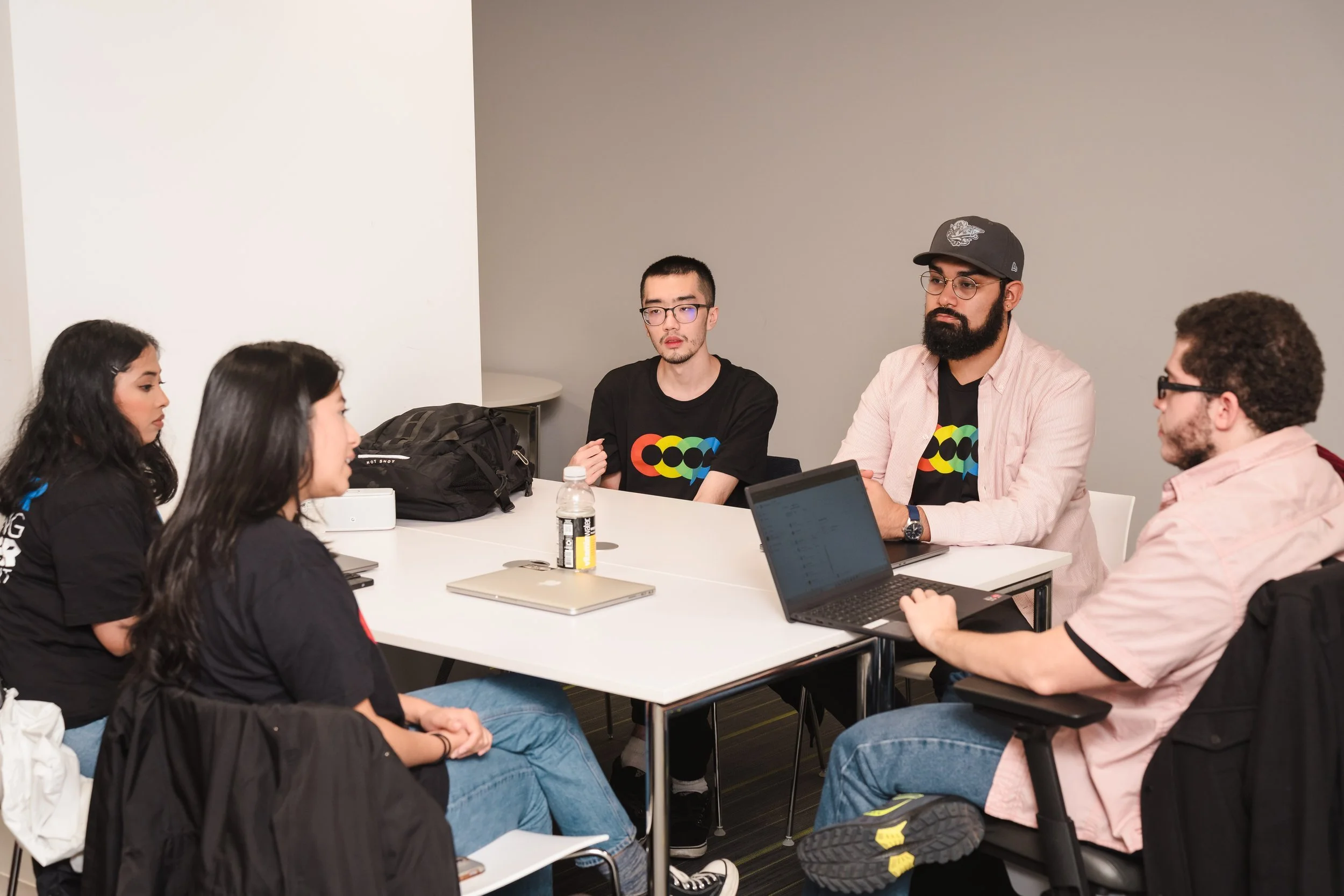 A group of six young people sitting around a white table in a room, participating in a meeting or discussion, with laptops, bottled water, and notebooks on the table.