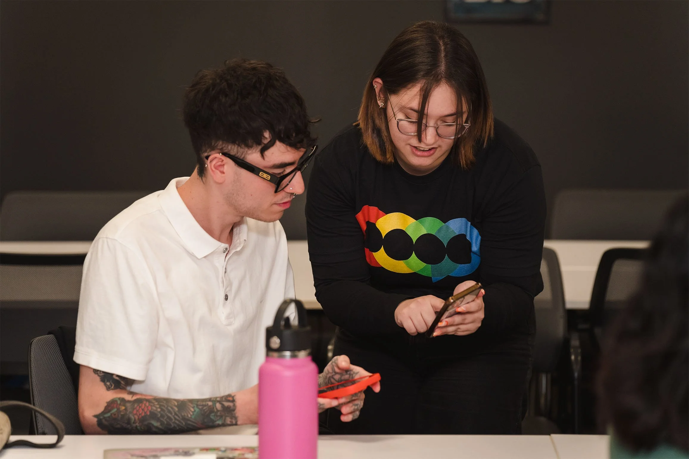 A man with tattoos and glasses sitting at a table, looking at his phone, with a woman standing next to him, showing him her phone. The woman has brown hair, glasses, and a black shirt with a colorful graphic. There is a pink water bottle in the foreground.