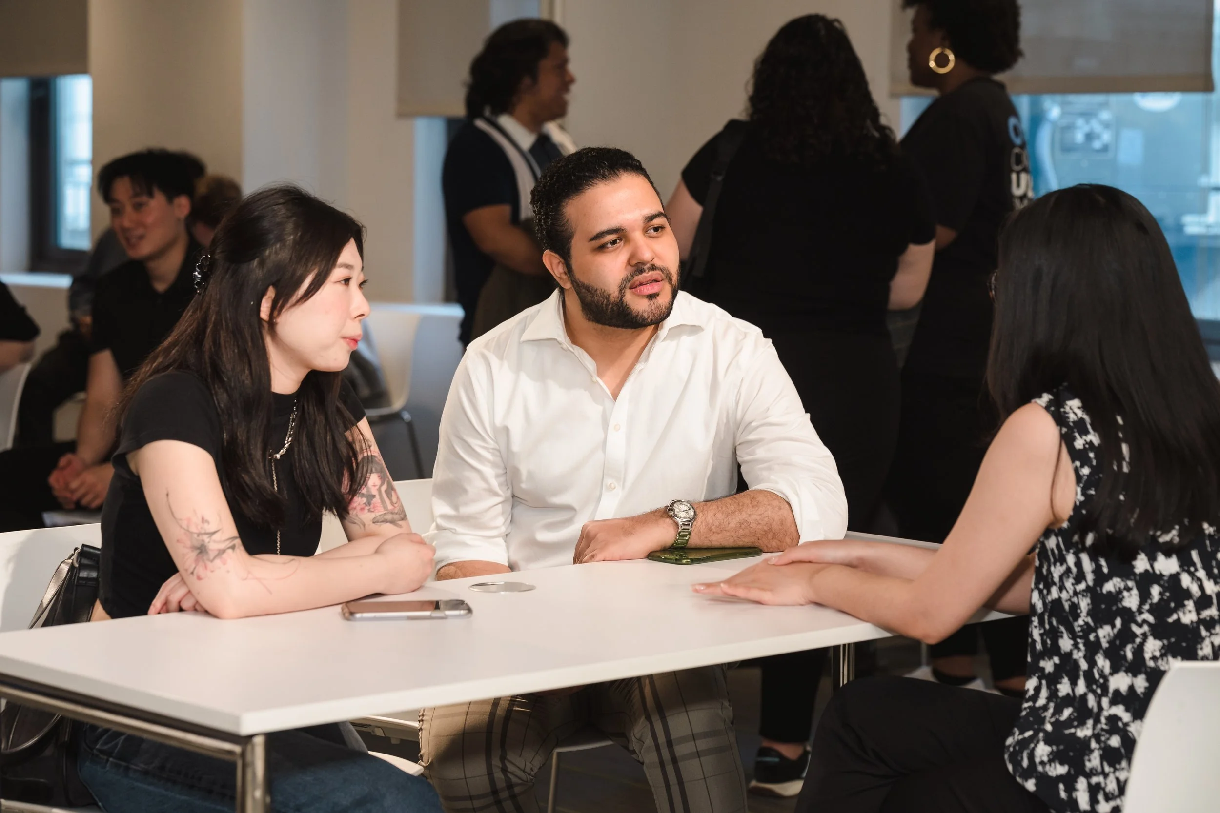 A diverse group of people gathered around a white table in a casual indoor setting, engaged in conversation.