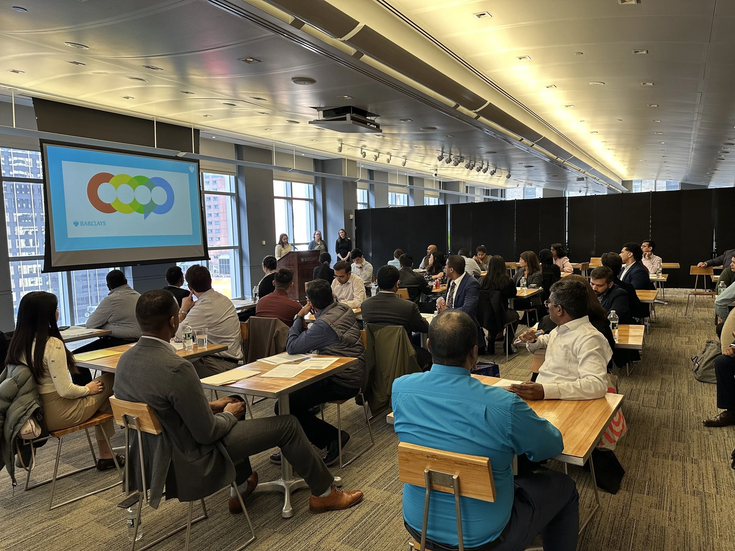 A large conference room filled with people seated at wooden tables listening to a presentation. Three women are standing near a podium in front of the room. A large screen displays a colorful logo with interconnected circles and the word 'Barclays'. The room has floor-to-ceiling windows with city buildings visible outside, and a modern ceiling with installed lighting.
