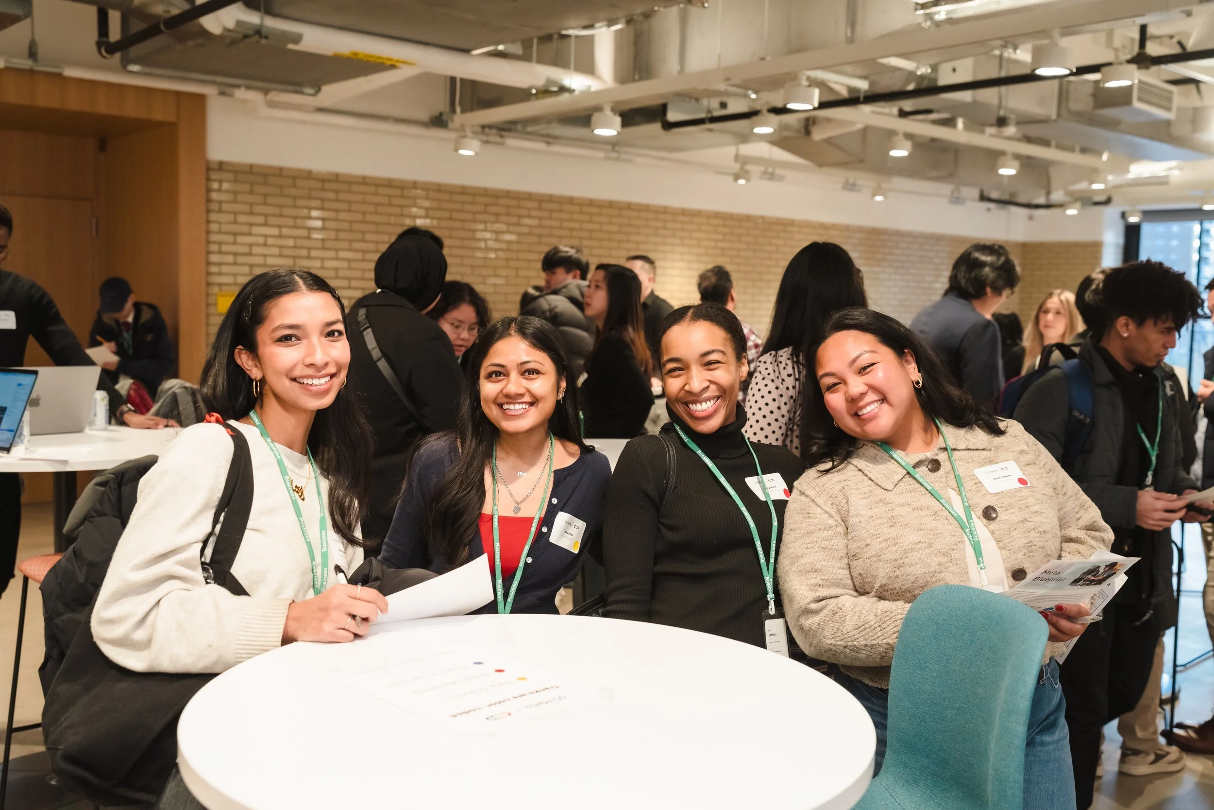 Four smiling women sitting at a table in a busy indoor conference or event space with many people in the background.
