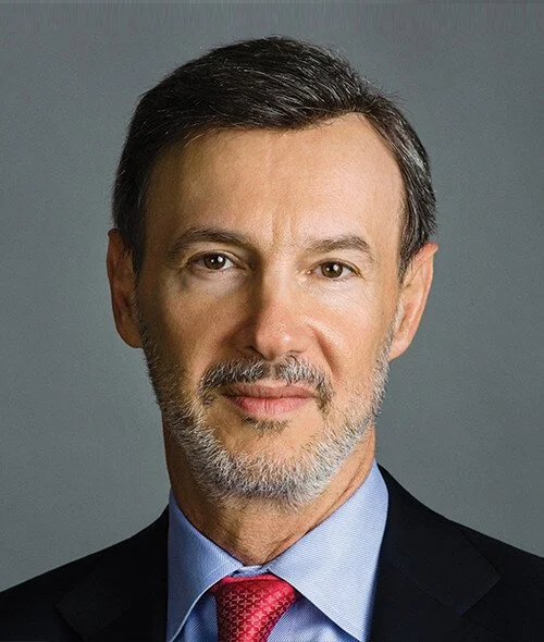 A professional headshot of a middle-aged man with short dark hair, a beard, wearing a dark suit, light blue shirt, and red tie, against a plain gray background.