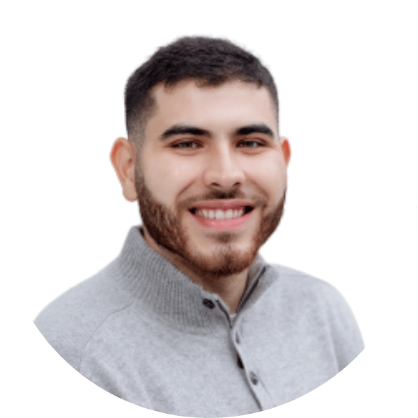 Portrait of a young man with short dark hair and a beard, smiling, wearing a light gray zip-up sweater, against a white background.