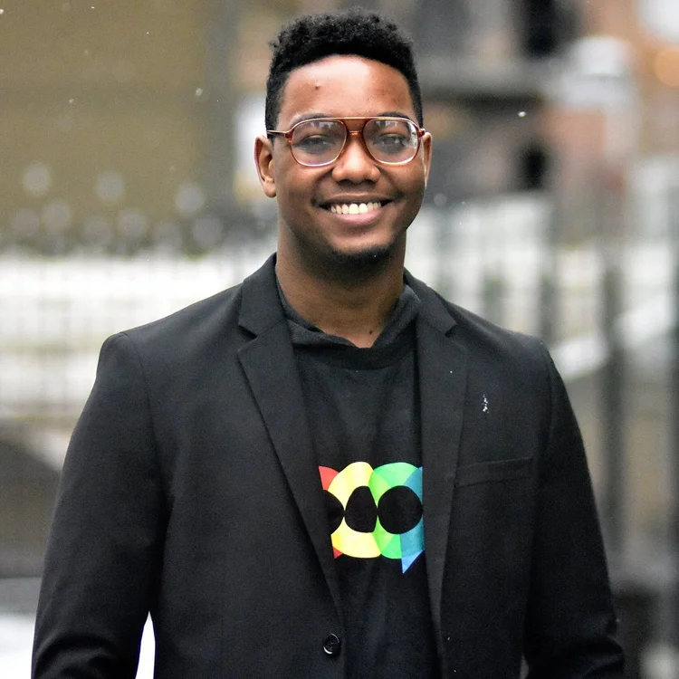 Smiling young man with glasses, wearing a black blazer over a black shirt with a colorful graphic, standing outdoors with blurred city background.