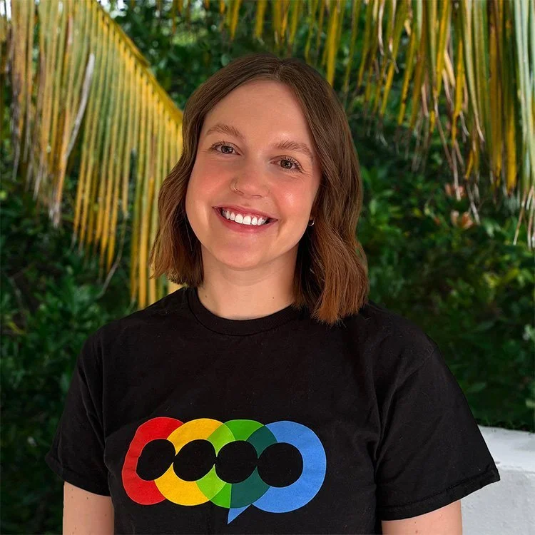 A young woman with shoulder-length brown hair, smiling, wearing a black T-shirt with a colorful rainbow-colored Olympic rings logo, standing outdoors with tropical greenery and palm leaves in the background.