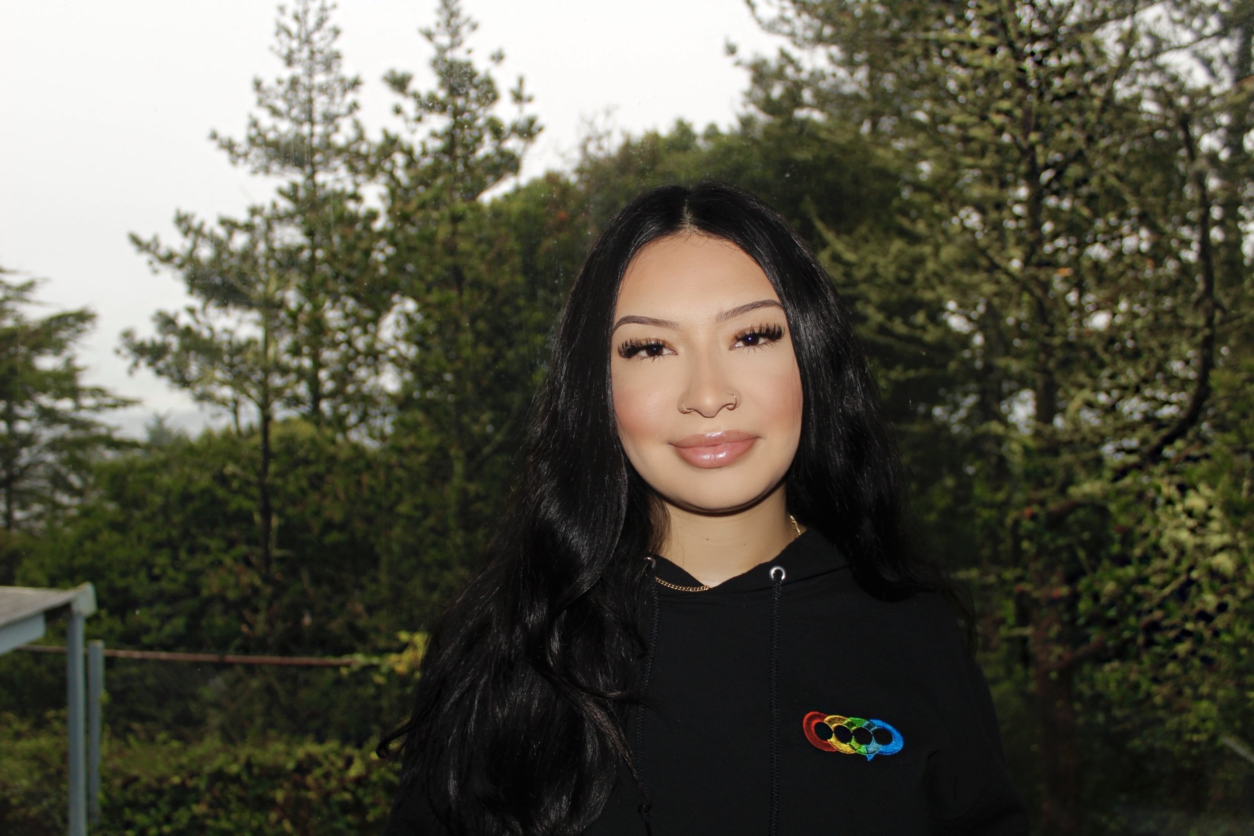 A woman with long black hair and a nose piercing, smiling outdoors with green trees in the background. She is wearing a black hoodie with a colorful rainbow logo.