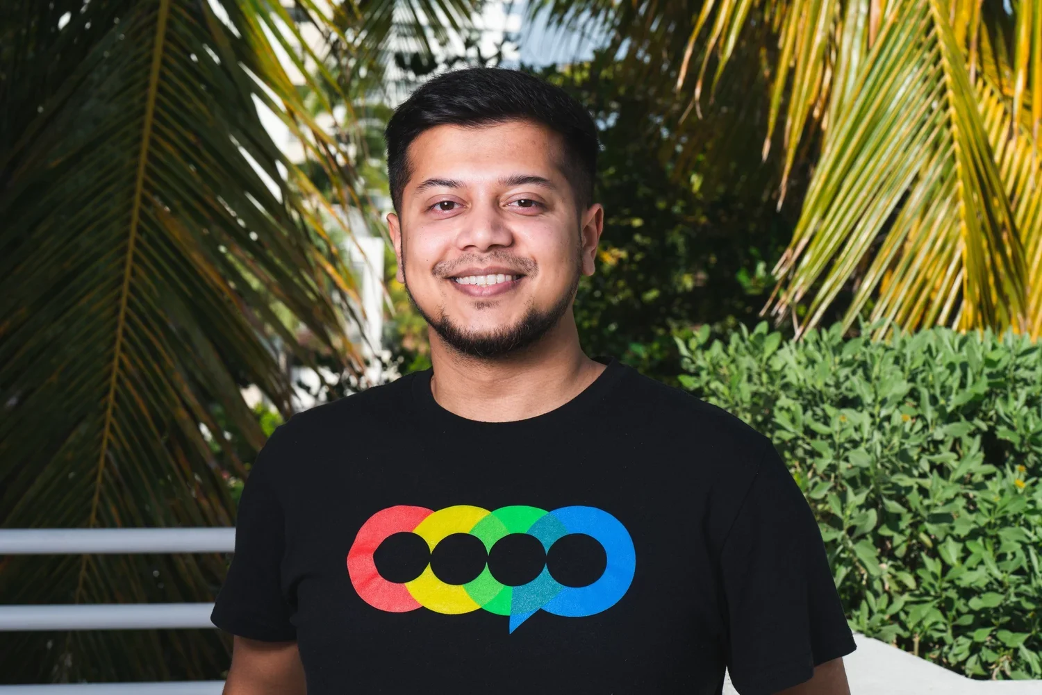 A smiling man with short dark hair and a goatee wearing a black T-shirt with colorful overlapping circles and text, standing outdoors with lush green plants and palm leaves in the background.