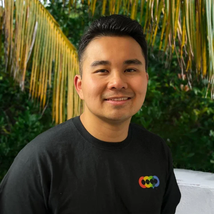 A smiling young man with short black hair, wearing a black t-shirt with a colorful logo, standing outdoors with green tropical plants and palm leaves in the background.