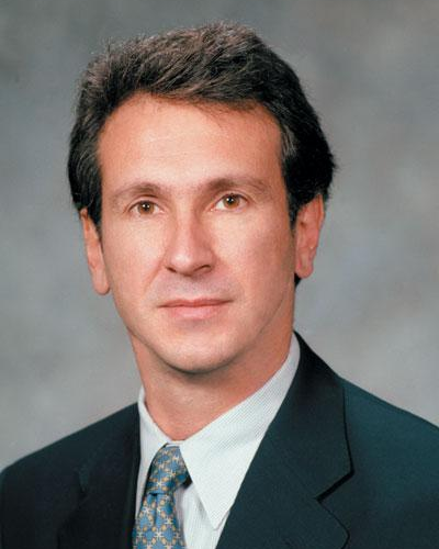A professional headshot of a man in a suit and tie with a neutral gray background.