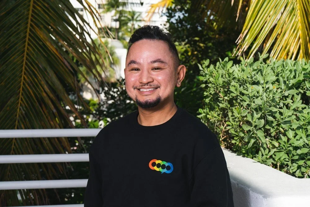 Smiling man with short dark hair and a goatee wearing a black sweatshirt with a colorful logo, standing outdoors with palm trees and green bushes in the background.