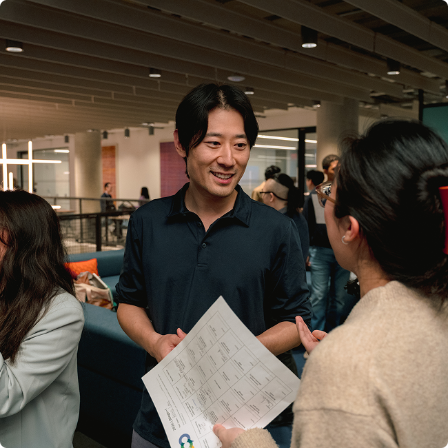 A young man smiling and holding a pamphlet, talking to a woman with glasses indoors at a busy event or conference.