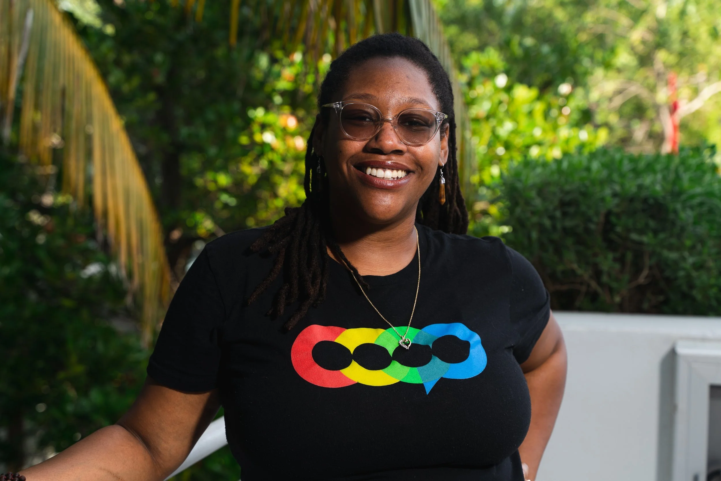 A smiling woman with glasses and dreadlocks wearing a black T-shirt with a colorful heart-shaped speaker design, standing outdoors in front of green foliage.