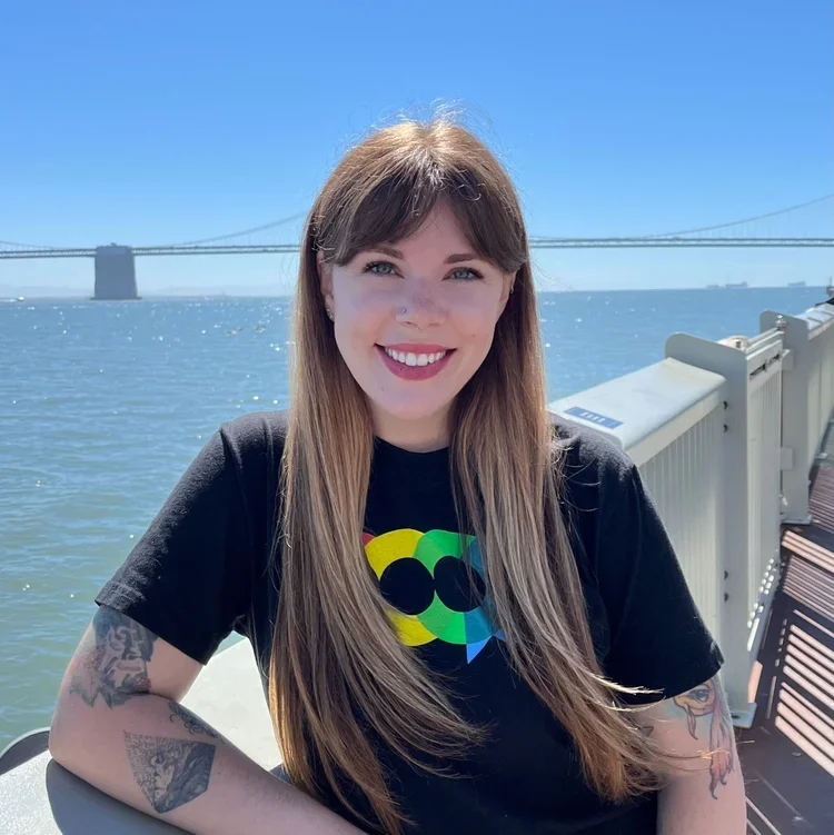 A smiling woman with long hair and tattoos on her arms, wearing a black T-shirt with a colorful design, sitting outdoors near a body of water with a bridge in the background.