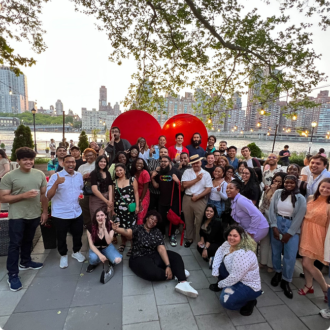 Large group of diverse people gathered outdoors in front of large red heart sculptures by a riverside, with city buildings in the background, during daytime.