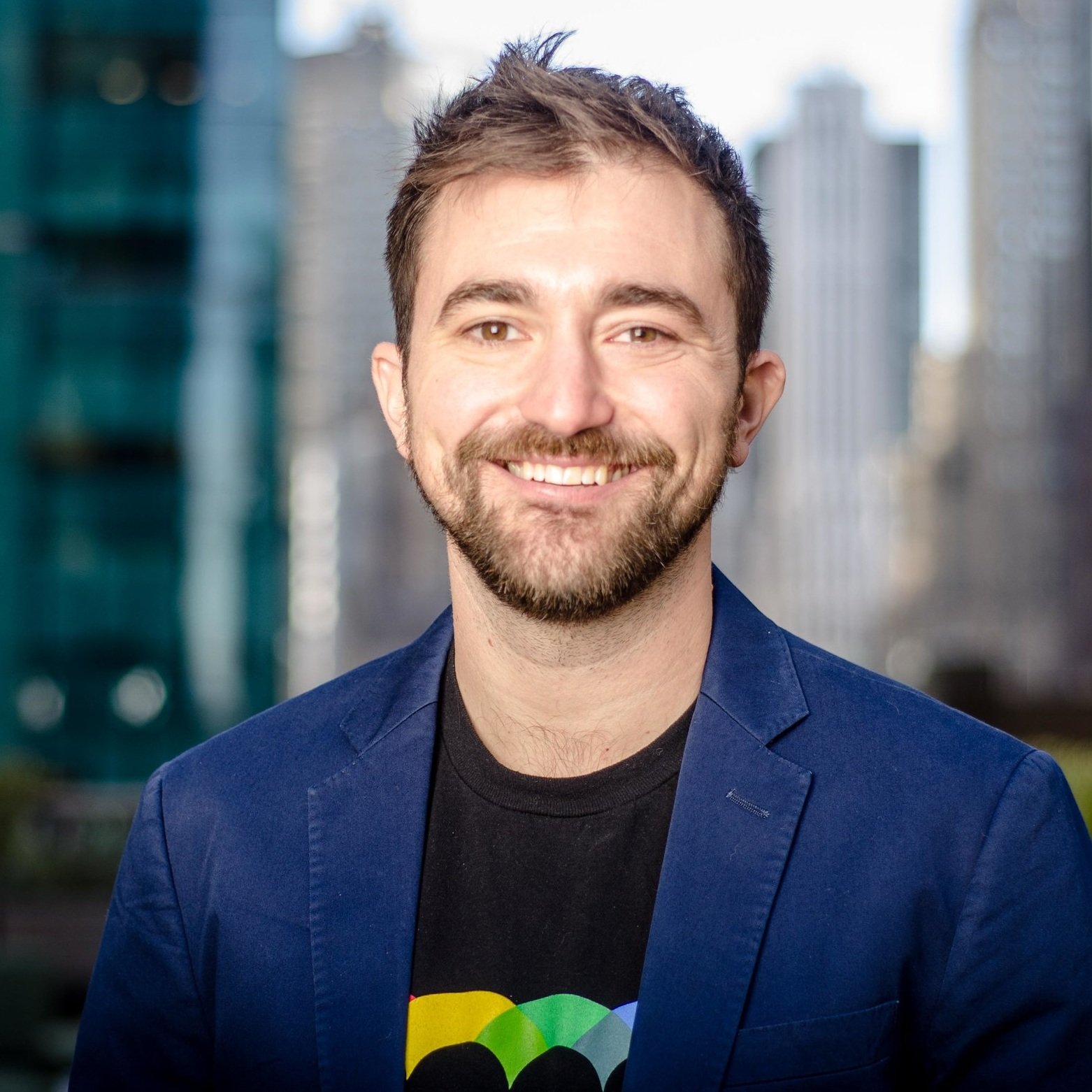 Headshot of a smiling man with beard and brown hair, wearing a blue blazer and a black t-shirt with a colorful heart design, in an urban cityscape background.