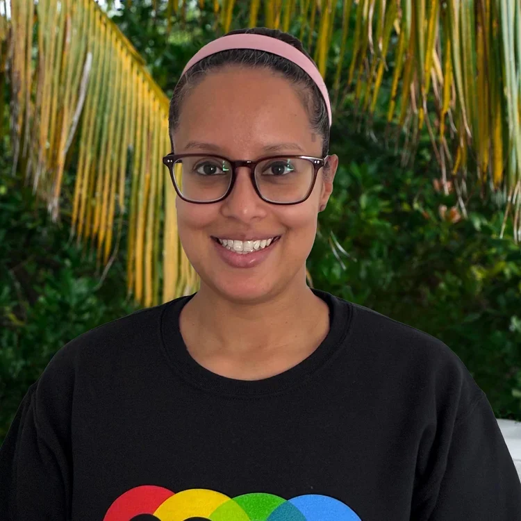 A woman wearing glasses and a black T-shirt with a rainbow-colored logo, smiling, with a pink headband, in front of a background of green tropical plants and palm leaves.