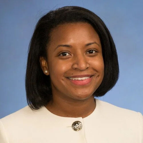 Headshot of a smiling African American woman with shoulder-length black hair, wearing a white blazer and pearl earrings, against a blue background.