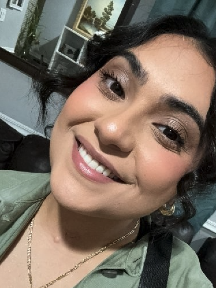 Close-up of a woman with dark hair, smiling, wearing makeup, a gold chain necklace, gold hoop earrings, and a green shirt.
