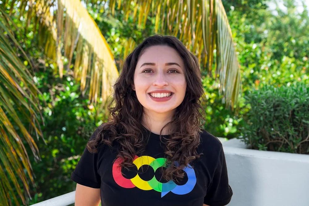 Young woman smiling outdoors with tropical plants and palm leaves in the background, wearing a black t-shirt with colorful Google logo.