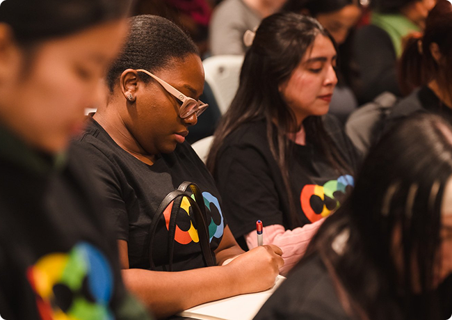 Group of diverse women sitting in a row, taking notes during an event or conference.