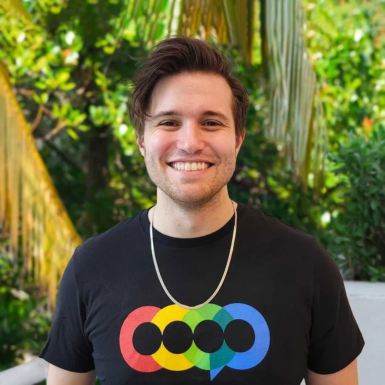 A smiling young man with brown hair, wearing a black t-shirt with a colorful overlapping circle design, standing outdoors with green trees and plants in the background.