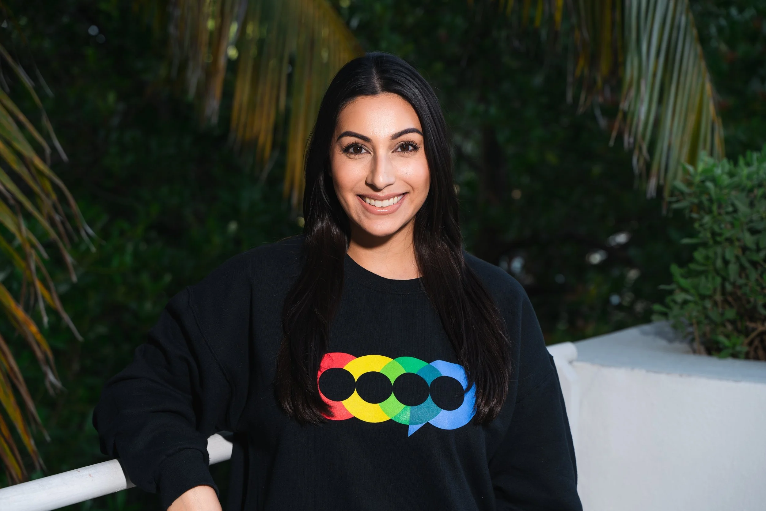 A young woman with dark hair and a bright smile, wearing a black sweatshirt with a colorful rainbow design, stands outdoors near a white railing with greenery in the background.