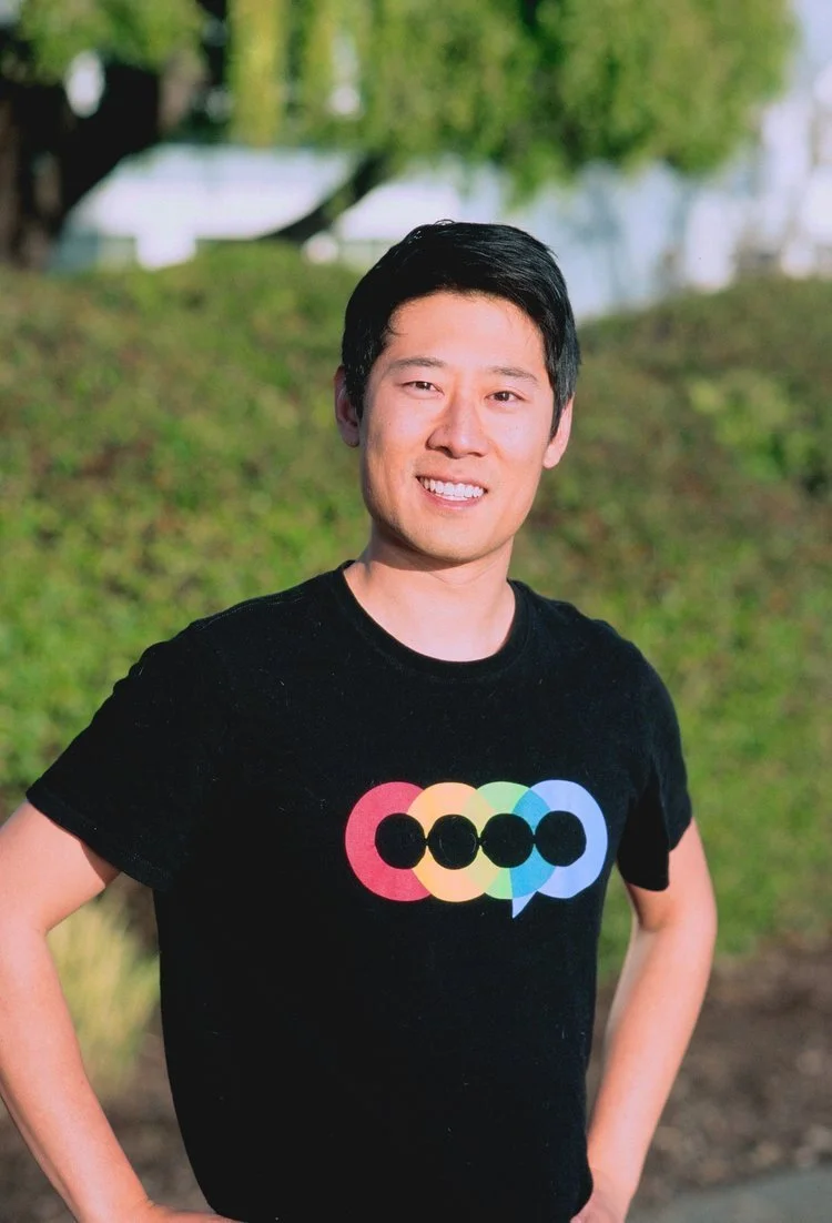 A young man with short black hair wearing a black T-shirt with a colorful logo, standing outdoors with grass and trees in the background.