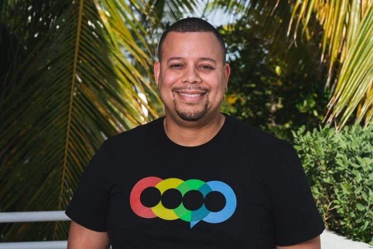 A smiling man standing outdoors with palm leaves and greenery in the background, wearing a black t-shirt with colorful overlapping circles and speech bubbles design.