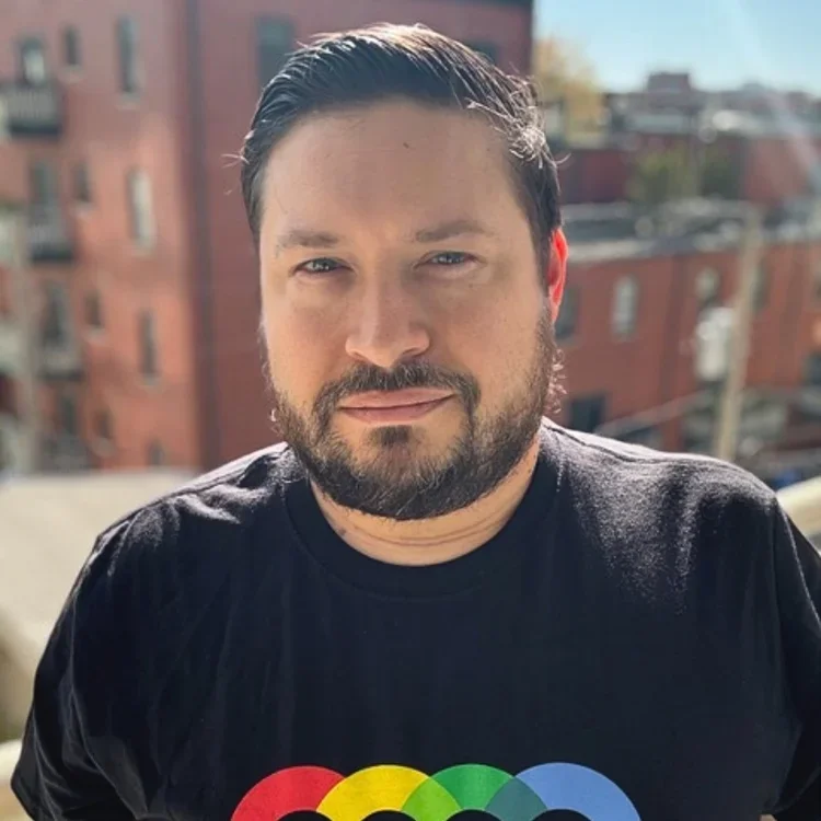 A man with dark hair and a beard taking a selfie outdoors on a sunny day, with a blurred cityscape in the background, wearing a black t-shirt with a colorful rainbow design.