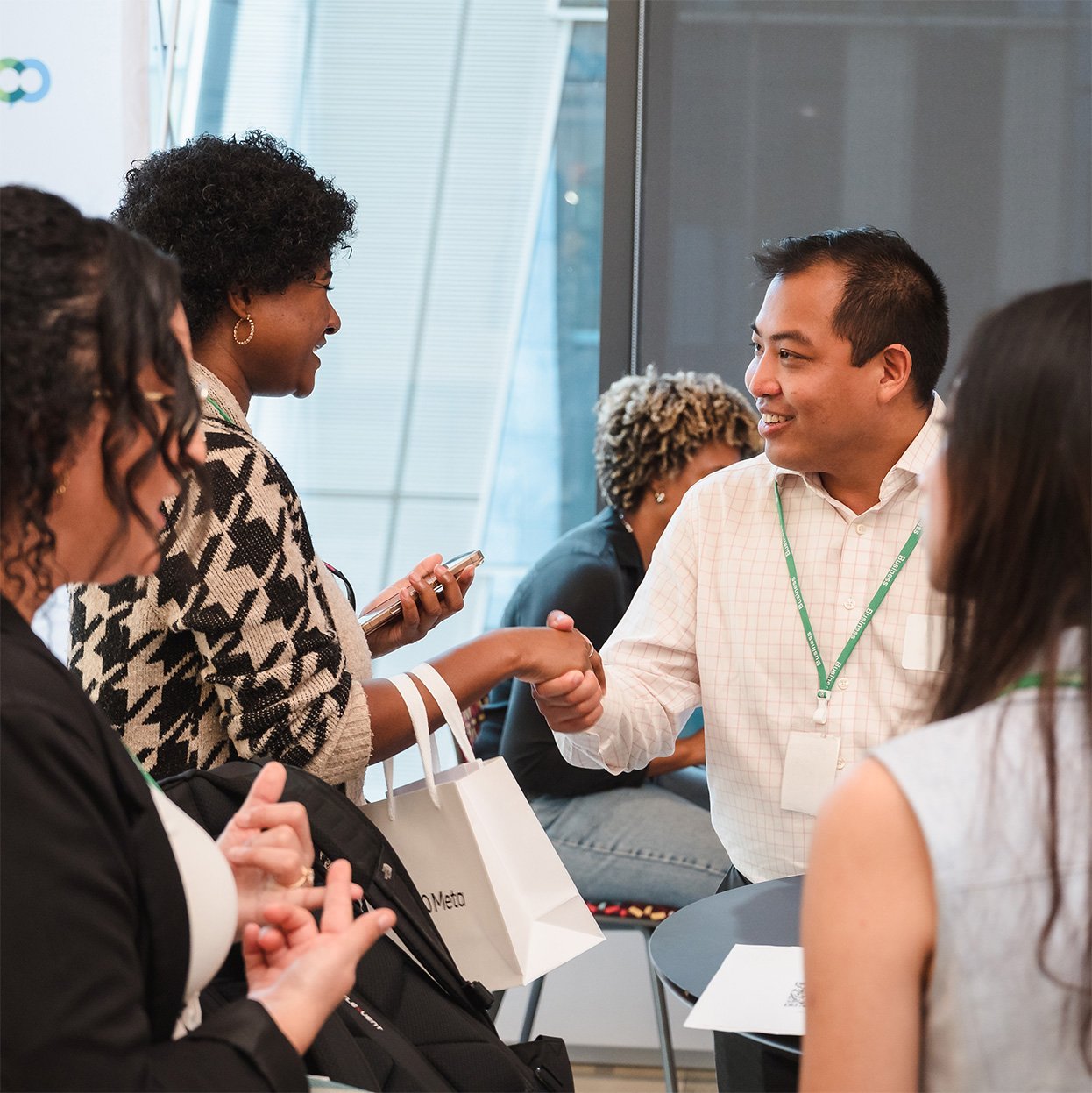 A diverse group of professionals at a conference, with one woman shaking hands with a man, all engaged in conversation near a large window.