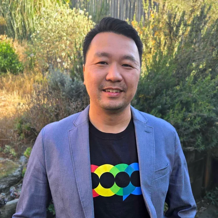 A smiling man in a gray blazer and black T-shirt with colorful circular logo, standing outdoors with green and yellow foliage in the background.