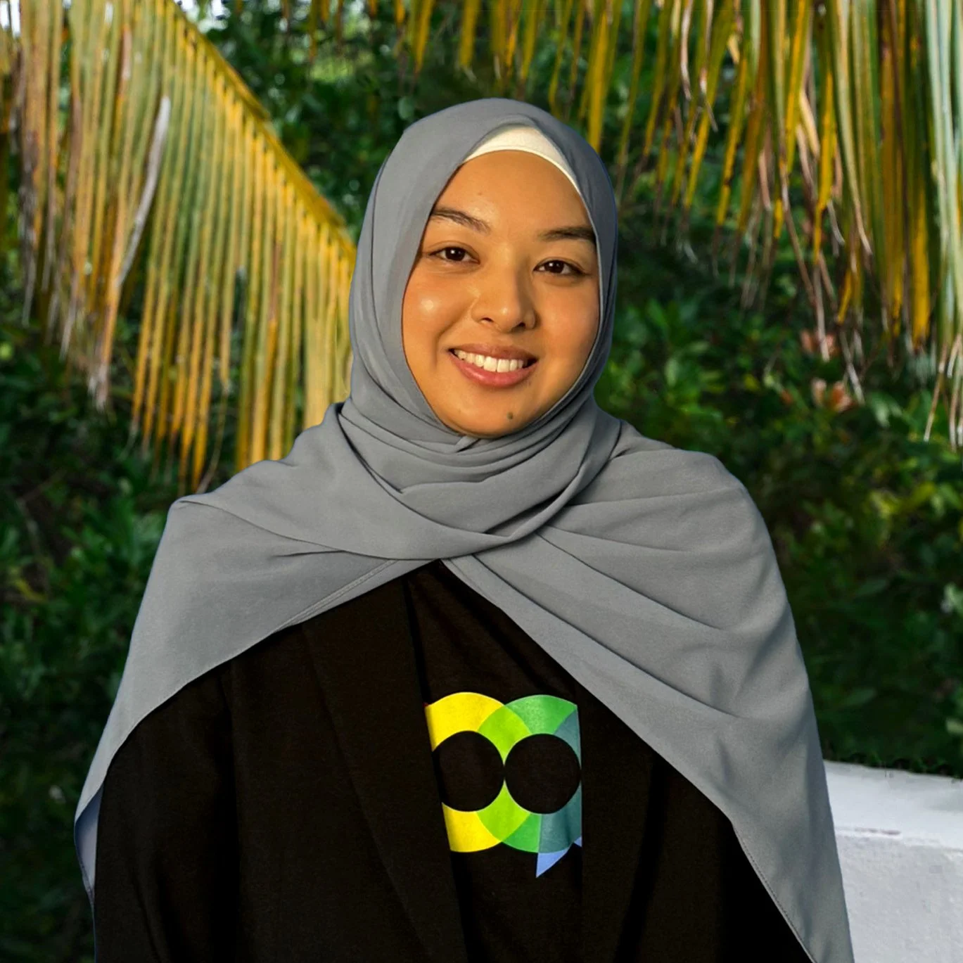 Portrait of a woman wearing a grey hijab, smiling, in a tropical outdoor setting with palm trees and foliage in the background.
