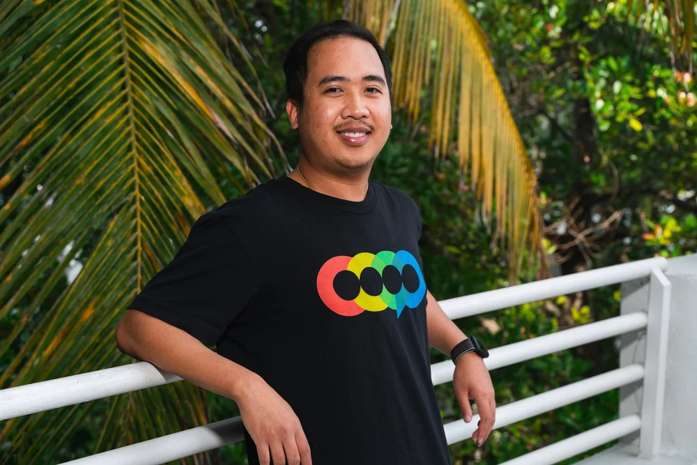A man leaning against a white railing outdoors with tropical palm trees in the background, wearing a black t-shirt with colorful speech bubble graphic.
